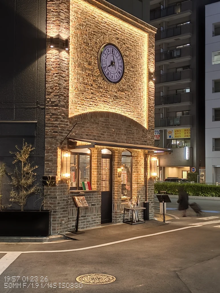 Nothing Phone (3a) Lite/カメラ:メイン(広角)2倍ズーム/焦点距離:50mm相当/内容:夜の街角で照明に浮かぶレンガ壁の建物と大きな時計が目立ち、歩道に人影も入るシーンを2倍で切り取った場面。