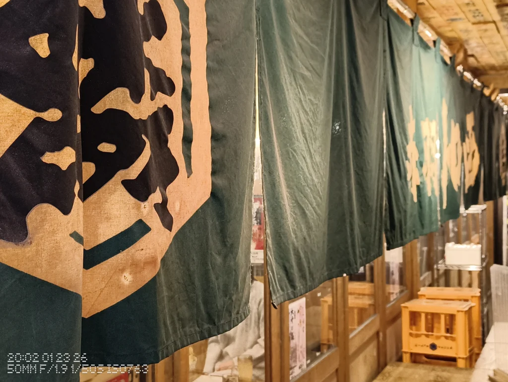 Nothing Phone (3a) Lite/カメラ:メイン(広角)2倍ズーム/焦点距離:50mm相当/内容:夜の店先に緑ののれんが大きく垂れ、暖色照明が反射する布地と入口周辺を2倍ズームで引き寄せた近景。