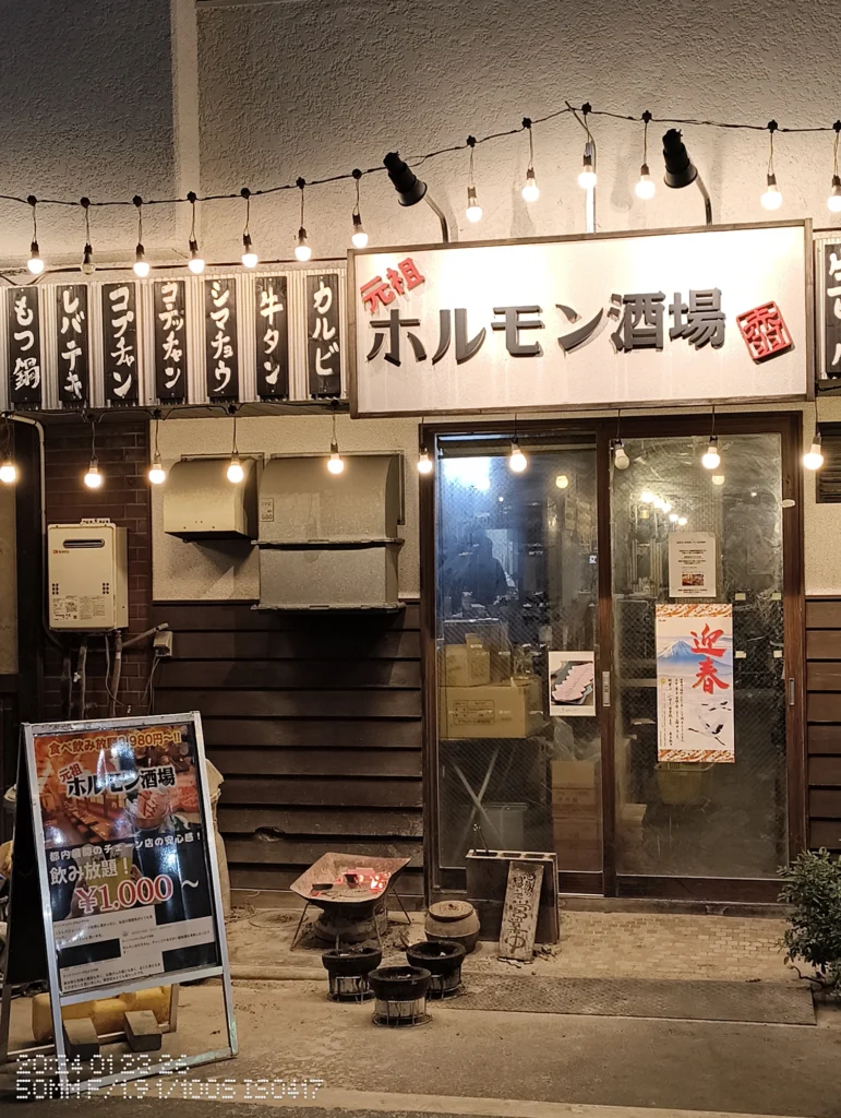 Nothing Phone (3a) Lite/カメラ:メイン(広角)2倍ズーム/焦点距離:50mm相当/内容:夜の飲食店前に店名看板と電球列が並び、メニュー看板や入口周りの光が強い路地の空気感を2倍で寄せた構図。