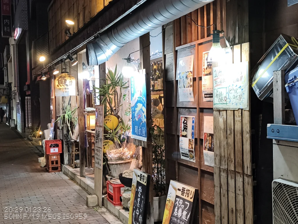 Nothing Phone (3a) Lite/カメラ:メイン(広角)2倍ズーム/焦点距離:50mm相当/内容:夜の歩道沿いに木壁の店構えとポスター、明るい裸電球が並び、奥へ続く飲食街を2倍ズームで引き寄せた夜景。