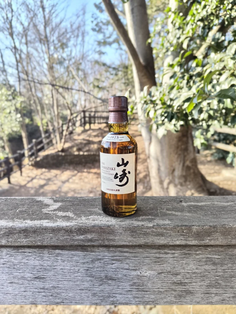 端末名:Nothing Phone (3a) Lite/カメラ:メイン(広角)ポートレート/焦点距離:確認不能(mm相当)mm相当/内容:木柵の上のウイスキーボトルを主題に背景ボケで公園の木々を溶かした昼のポートレート、透明感と立体感を強調