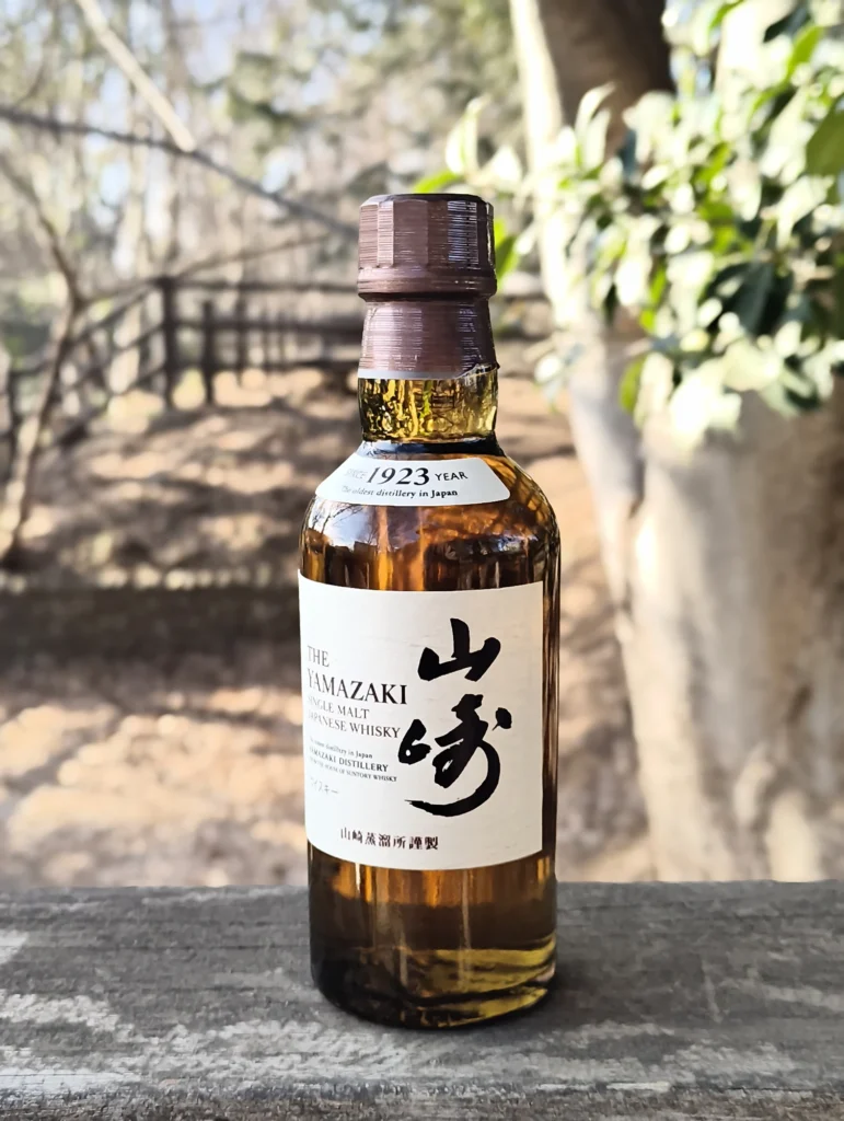 端末名:Nothing Phone (3a) Lite/カメラ:メイン(広角)ポートレート/焦点距離:確認不能(mm相当)mm相当/内容:ウイスキーボトルのラベルを中心に背景ボケで林の景色を溶かした昼のポートレート、反射と質感で立体感を演出