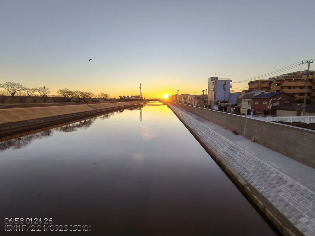 Nothing Phone (3a) Lite/超広角(15mm相当)、運河の水面に朝日が映り、両岸の護岸と住宅が一点へ収束する早朝の景、空のグラデーション