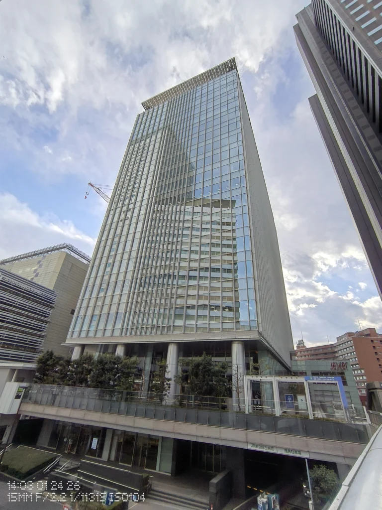 Nothing Phone (3a) Lite/超広角(15mm相当)、ガラス張りの高層ビルを見上げ、反射する空と周囲の建物が歪み少なく写る、上部に薄雲