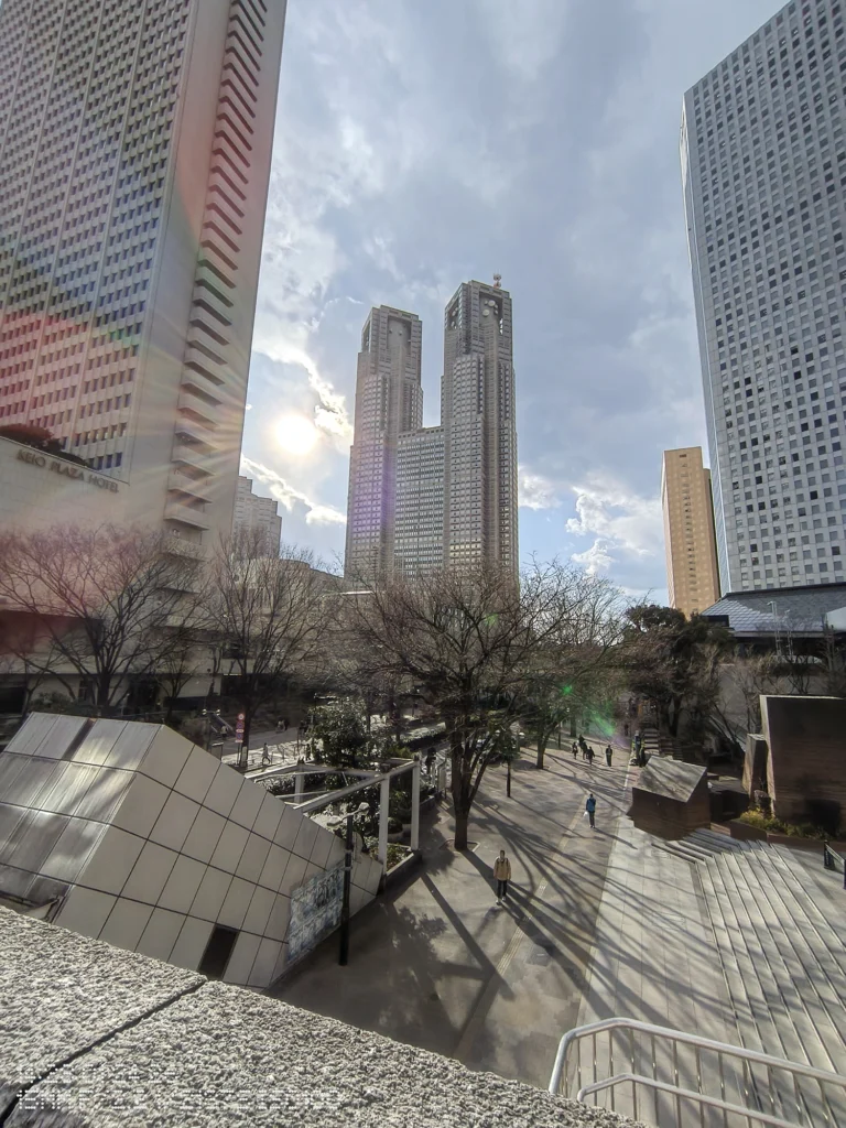 Nothing Phone (3a) Lite/超広角(15mm相当)、高層ビル群の間に低い太陽が入り、フレアと長い影が伸びる夕方の広場、空に薄雲が流れる