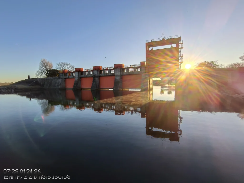 Nothing Phone (3a) Lite/超広角(15mm相当)、赤い水門と水面の反射、右端の低い太陽で光芒とフレアが出た河川施設、水面は鏡のよう
