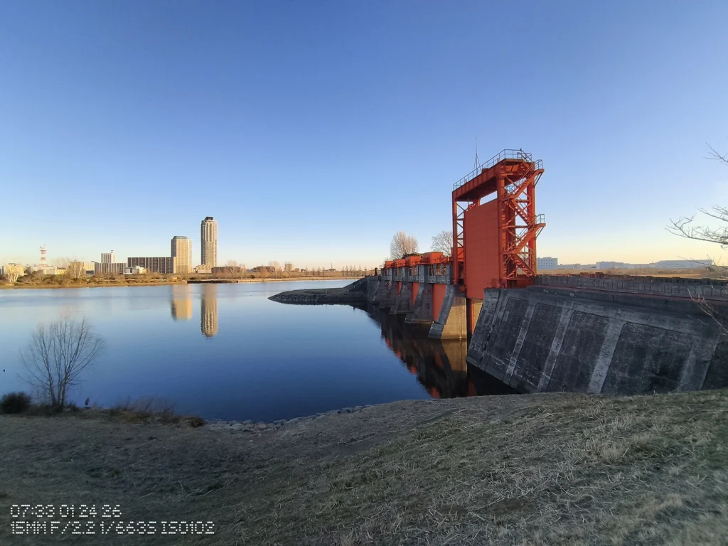 Nothing Phone (3a) Lite/超広角(15mm相当)、湖面と赤い水門を右に、左奥に高層ビル群が映り込む冬の晴天の広景、水際の護岸も写る