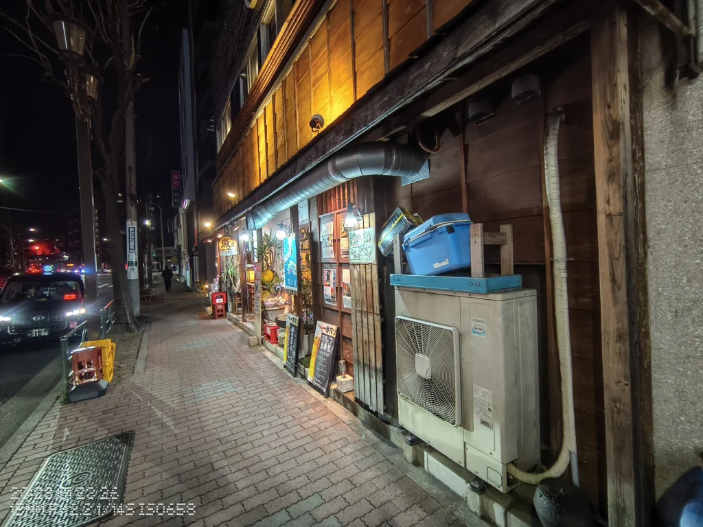Nothing Phone (3a) Lite/カメラ:超広角/焦点距離:15mm相当/内容:夜の歩道に木造外壁と室外機が迫り、店先の暖色照明と車道の街灯が奥へ伸びる夜景