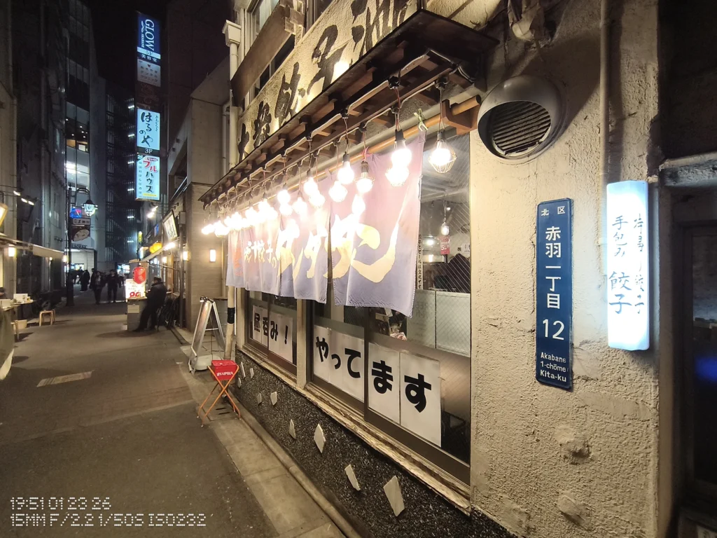 Nothing Phone (3a) Lite/カメラ:超広角/焦点距離:15mm相当/内容:夜の飲食店の軒先に裸電球が並び、のれんと店内光が路地を照らす温かい夜景