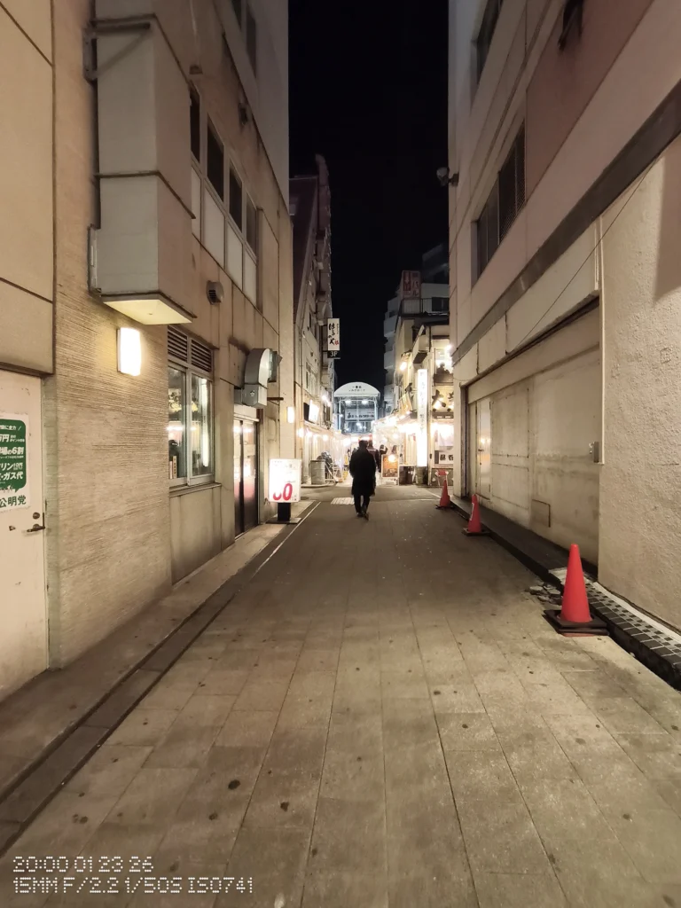 Nothing Phone (3a) Lite/カメラ:超広角/焦点距離:15mm相当/内容:夜の細い路地に店の看板と街灯が続き、歩く人のシルエットと奥行きが強調された夜景