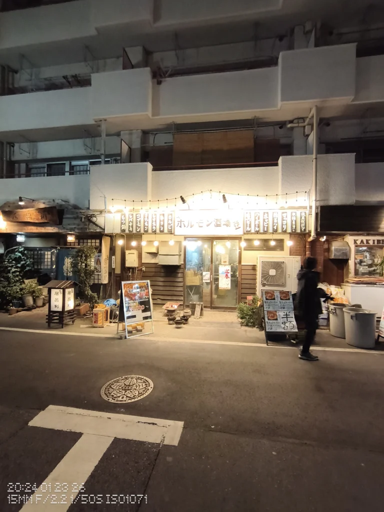 Nothing Phone (3a) Lite/カメラ:超広角/焦点距離:15mm相当/内容:夜の飲食店の正面に明るい看板と電球が並び、建物前を歩く人がブレて写る夜景