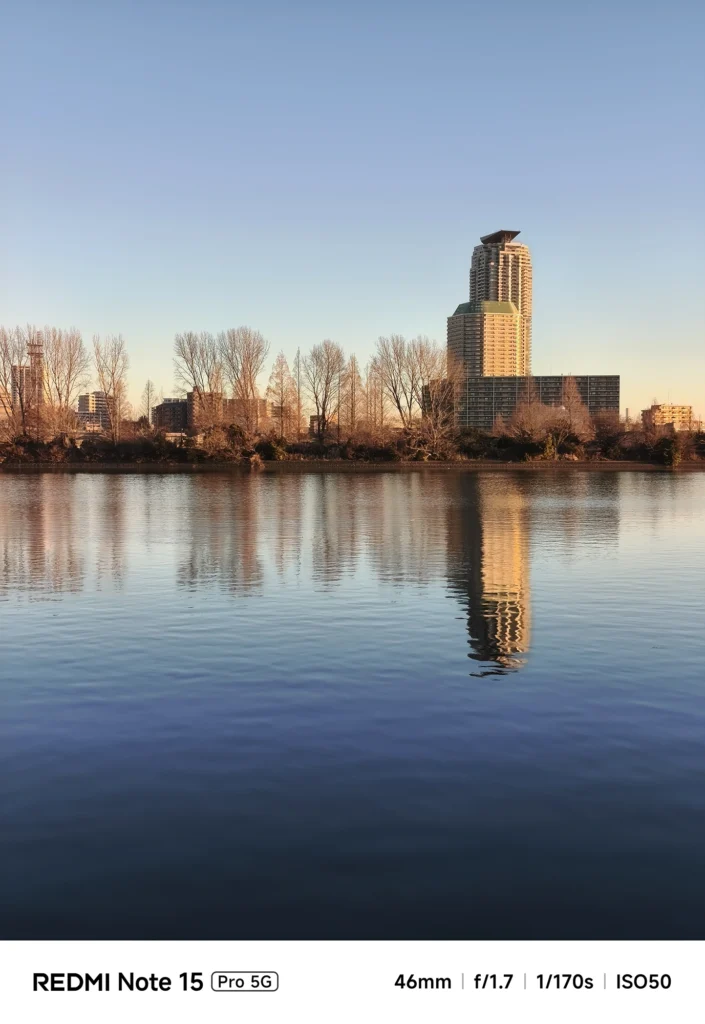 穏やかな川面に反射する都市のビル群と冬の木々。静寂を感じる水辺の風景。Redmi Note 15 Pro 5Gで撮影。