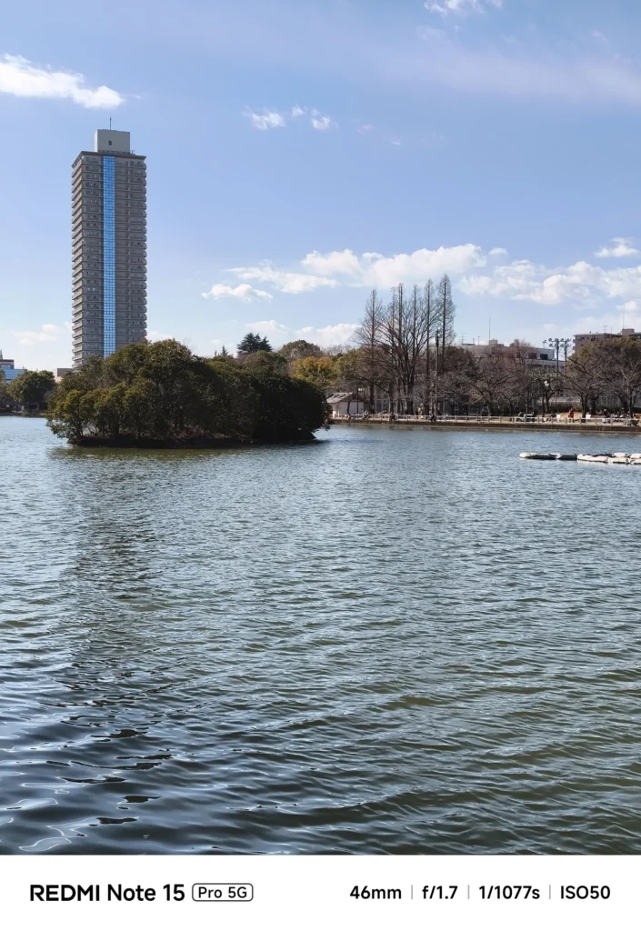青空の下、波立つ水面越しに望む公園の島と高層マンション。開放感のある都会のオアシス。Redmi Note 15 Pro 5Gで撮影。