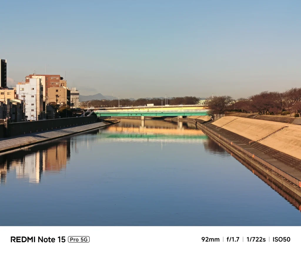 穏やかな川面に映る橋と街並み。ズームによる圧縮効果で静寂な川の広がりを強調した風景。Redmi Note 15 Pro 5G(92mm)で撮影。