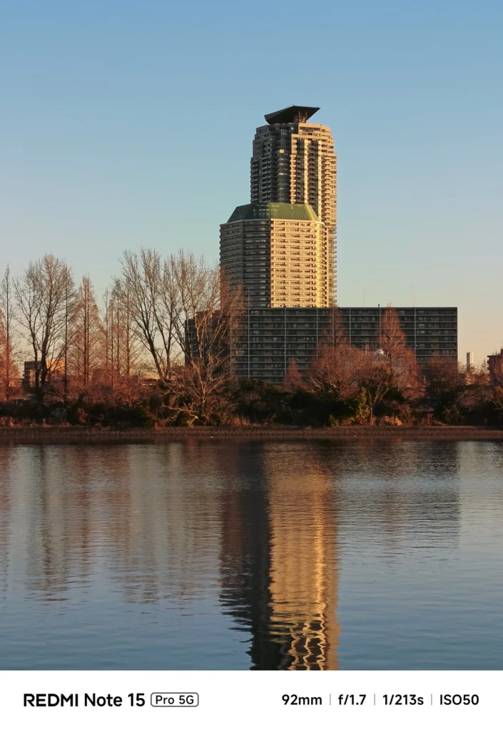 夕暮れ時の黄金色の光に照らされた高層ビルと、水面に長く伸びるその反射。Redmi Note 15 Pro 5G(92mm)で撮影。
