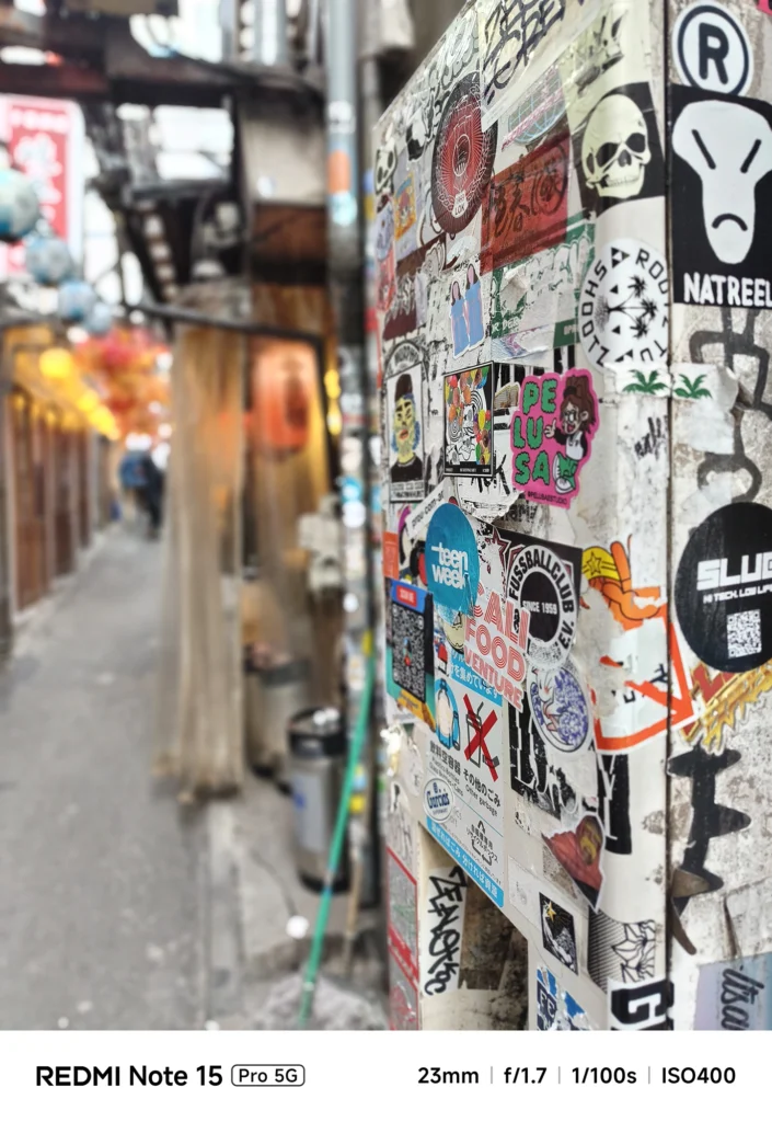 街角の分電盤に貼られたストリートアート風ステッカーの接写。背景の路地を大きくぼかした構図。Redmi Note 15 Pro 5G(ポートレートモード)で撮影。