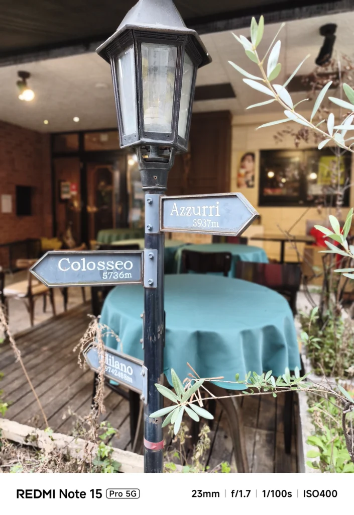カフェのテラスに立つ、イタリアの都市(Azzurri, Milano等)を指す方向サイン。Redmi Note 15 Pro 5G(ポートレートモード)で撮影。