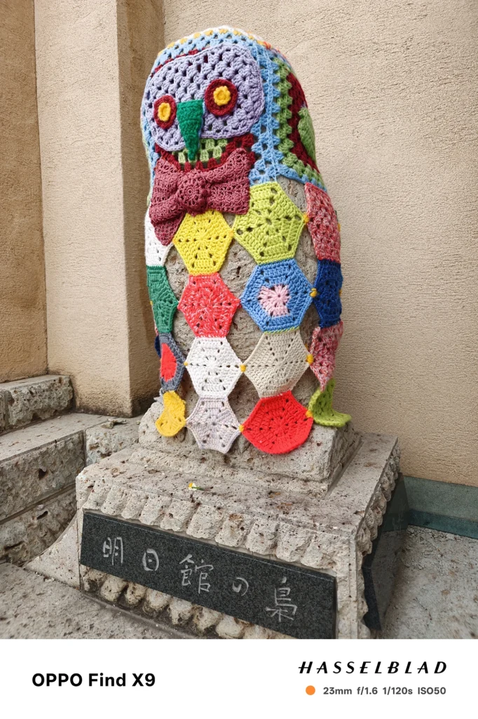 端末名：Find X9／カメラ：メイン（広角）／焦点距離：23mm相当／内容：多色の毛糸で編まれたフクロウ装飾が石台に据えられ、壁面の質感と併せて見える屋外の展示