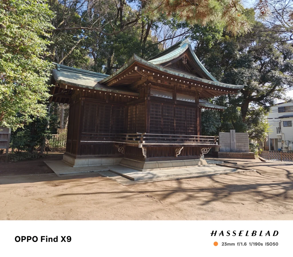 端末名：Find X9／カメラ：メイン（広角）／焦点距離：23mm相当／内容：木造の社殿を斜めから捉え、境内の木立と地面の影が伸びる昼の神社風景を広く写した構図