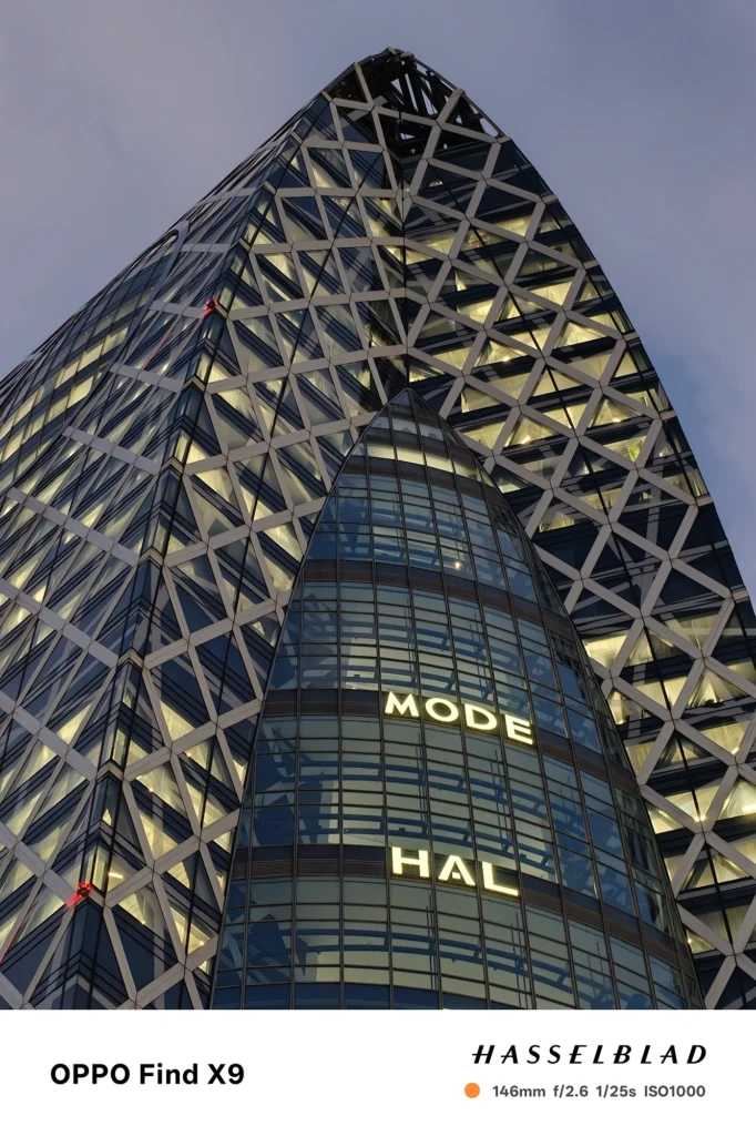 端末名：Find X9／カメラ：望遠／焦点距離：146mm相当／内容：格子外装の高層ビルを望遠で引き寄せ、窓明かりと発光文字が夜景の圧縮感を強める静かな都市の雰囲気