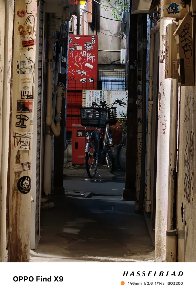 端末名：Find X9／カメラ：望遠／焦点距離：146mm相当／内容：路地奥の自転車と赤い設備、壁面のステッカーや配線を望遠で圧縮し、点在する光源が奥行きを作る夜景撮影