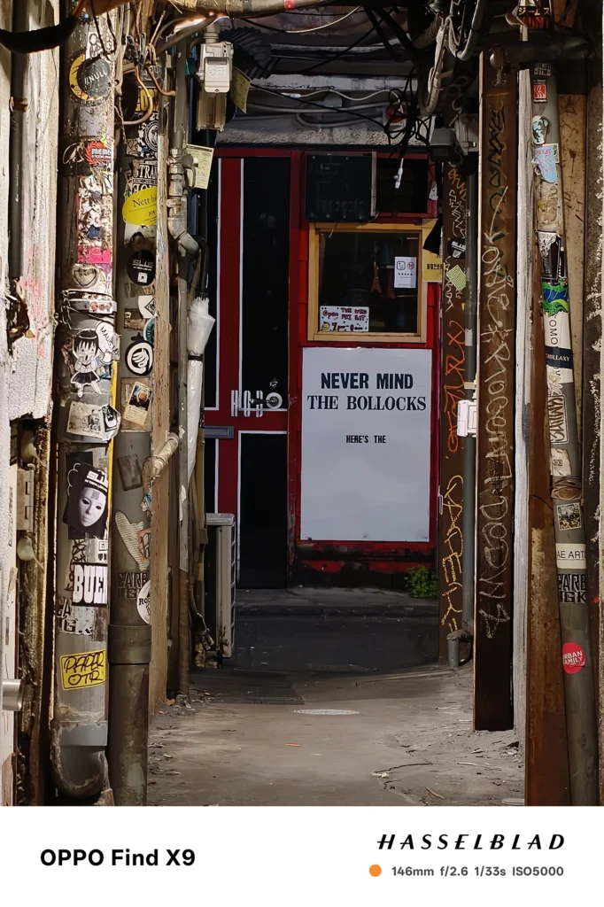 端末名：Find X9／カメラ：望遠／焦点距離：146mm相当／内容：路地の扉と英字の大きな看板、周囲の貼り紙や配線を望遠で引き寄せ、夜の光源が圧縮感と退廃的な空気を強める