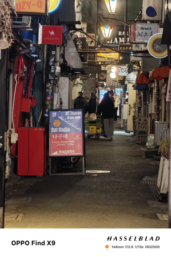 端末名：Find X9／カメラ：望遠／焦点距離：146mm相当／内容：細い路地に並ぶ看板と照明、歩行者のシルエットを望遠で引き寄せ、雑多な光源が圧縮効果で奥へ続く夜景の雰囲気