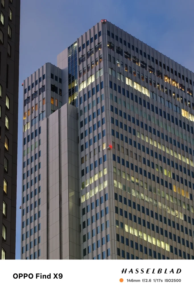 端末名：Find X9／カメラ：望遠／焦点距離：146mm相当／内容：高層ビルの外壁と点在する窓明かりを望遠で圧縮し、薄暮の青空と直線的な構造が夜景の立体感を作る