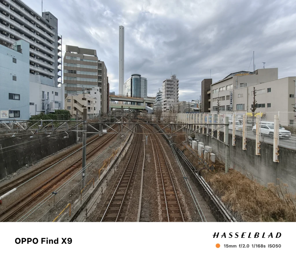 Find X9／カメラ：超広角／焦点距離：15mm相当／内容：都市部の線路を跨ぐ位置から複数の線路と高層建物、曇り空を広く写した風景