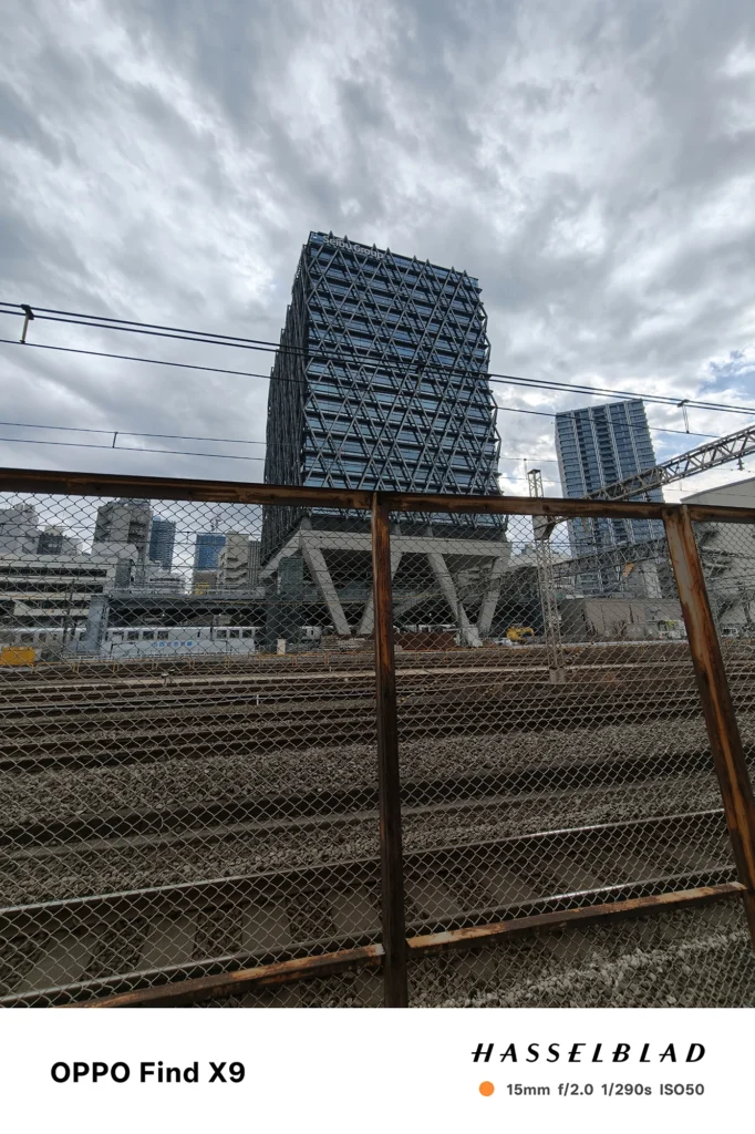 端末名：Find X9／カメラ：超広角／焦点距離：15mm相当／内容：線路脇のフェンス越しに高層ビルと架線を広角で捉え、曇り空の下で建物の格子構造と鉄道設備が見える昼間の街並み