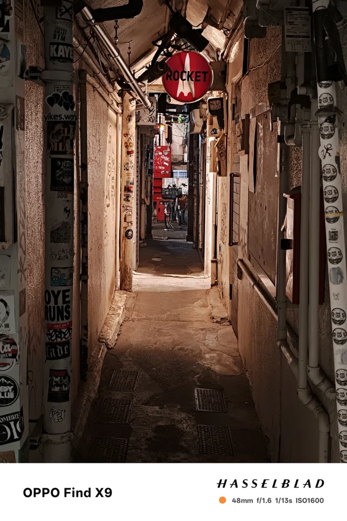 端末名：Find X9／カメラ：メイン（広角）2倍ズーム／焦点距離：48mm相当／内容：看板が連なる路地を2倍ズームで切り取り、自転車の並びと店先の照明、奥の人影が混在光で浮かぶ夜景