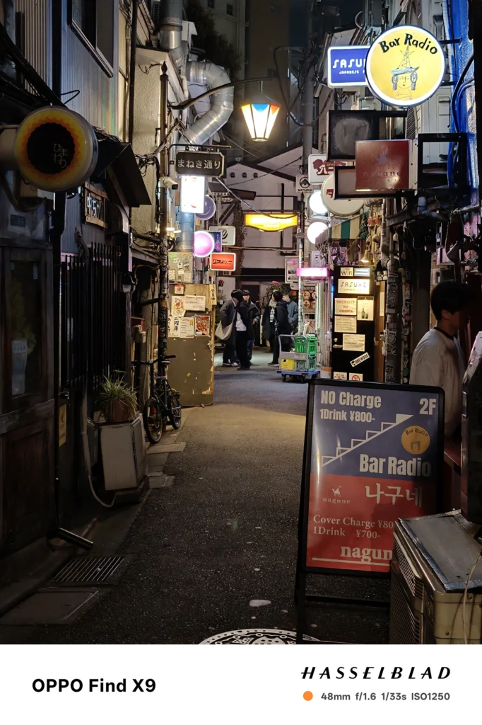 端末名：Find X9／カメラ：メイン（広角）2倍ズーム／焦点距離：48mm相当／内容：Bar Radioの看板と立て看板が見える路地を2倍ズームで切り取り、店灯りと点光源の連なりが奥へ続く夜景