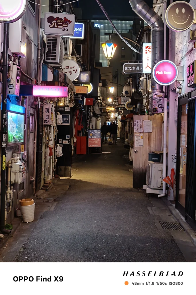 端末名：Find X9／カメラ：メイン（広角）2倍ズーム／焦点距離：48mm相当／内容：ネオン看板が密集する細い路地を2倍ズームで引き寄せ、発光サインの点光源と奥の人影が連なる繁華街の夜景