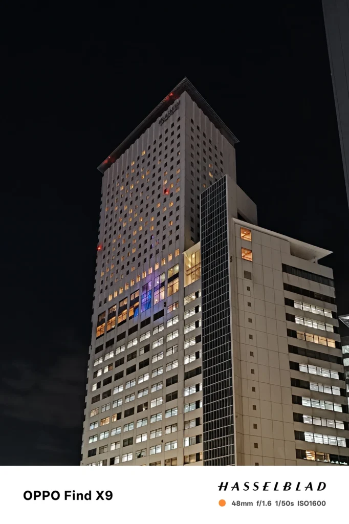 端末名：Find X9／カメラ：メイン（広角）2倍ズーム／焦点距離：48mm相当／内容：白い高層ビルを2倍ズームで見上げ、窓明かりの点光源と黒い夜空のコントラストが強い都市夜景