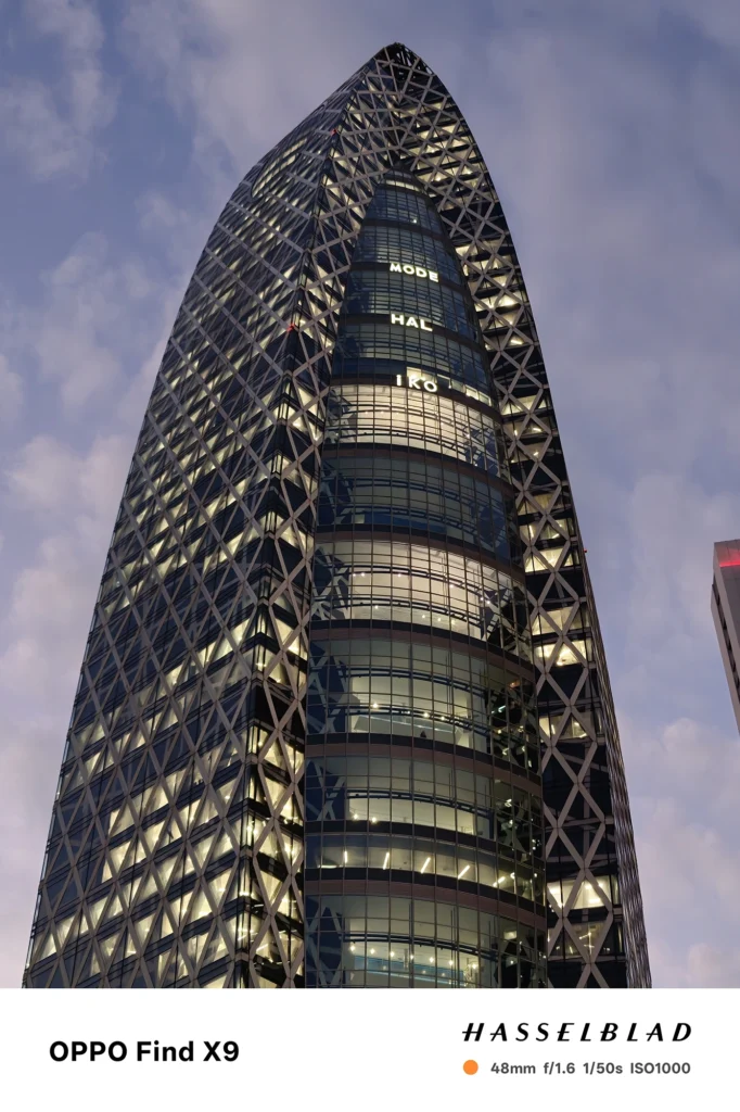 端末名：Find X9／カメラ：メイン（広角）2倍ズーム／焦点距離：48mm相当／内容：格子状外装の高層ビルを見上げ、窓明かりの点光源と薄暮の雲が重なる夜景を2倍ズームで引き寄せた構図