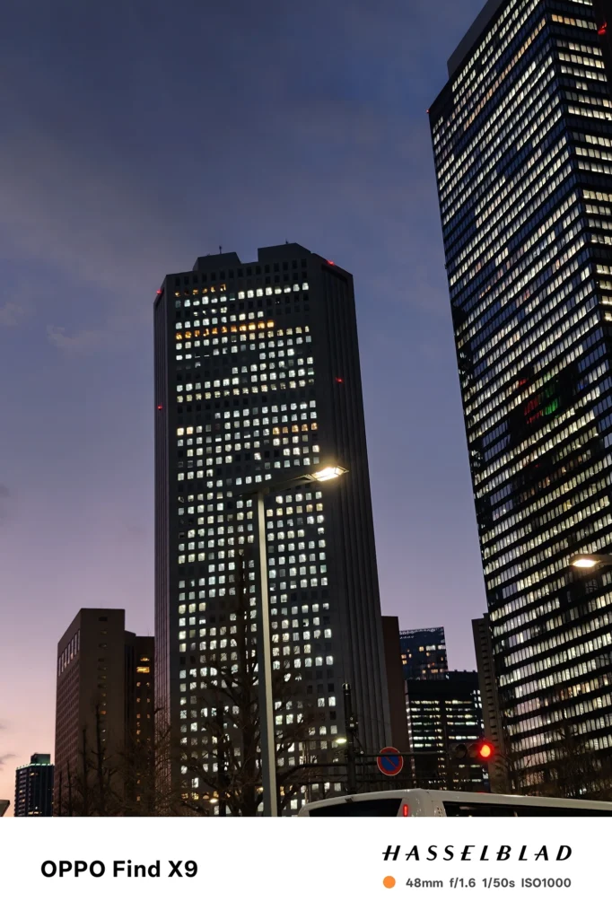 端末名：Find X9／カメラ：メイン（広角）2倍ズーム／焦点距離：48mm相当／内容：高層ビル群と街灯を2倍ズームで切り取り、窓明かりの点光源と夕暮れの青紫の空が同居する都市夜景