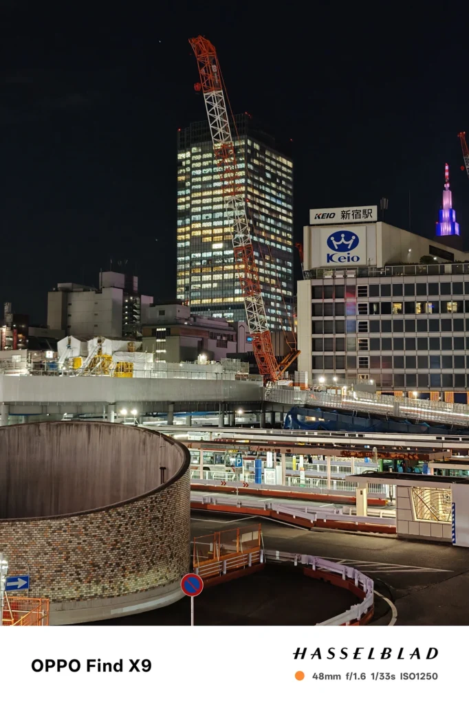 端末名：Find X9／カメラ：メイン（広角）2倍ズーム／焦点距離：48mm相当／内容：クレーンとビル群を2倍ズームで切り取り、Keio新宿駅の看板と窓明かりの点光源が浮かぶ工事現場周辺の夜景