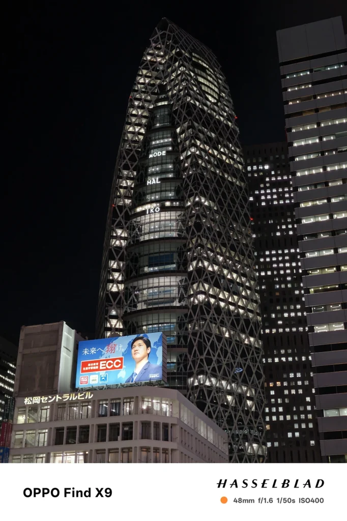 端末名：Find X9／カメラ：メイン（広角）2倍ズーム／焦点距離：48mm相当／内容：ネオン看板が並ぶ大通りを2倍ズームで切り取り、車列の灯りと発光看板の点光源が密集する繁華街の夜景