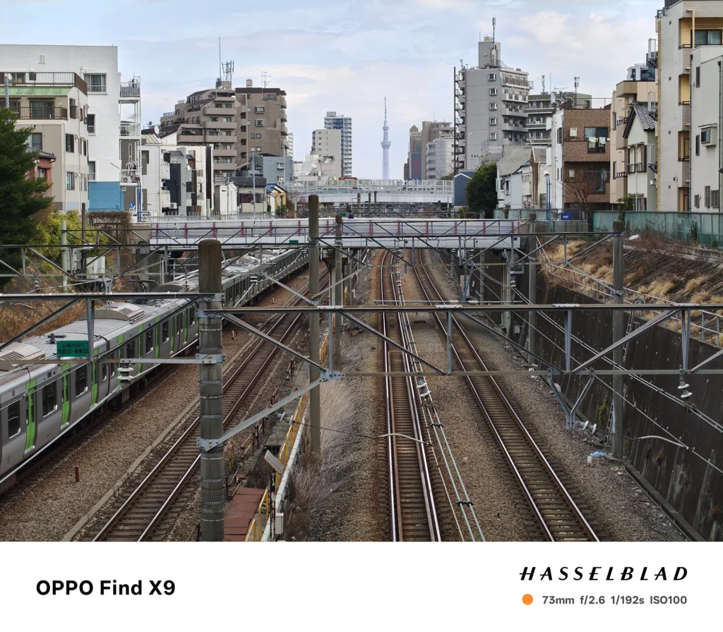 OPPO Find X9で撮影した73mm中望遠の鉄道風景。複数の線路と架線、走行中の列車、遠方の街並みを中央に捉えている。