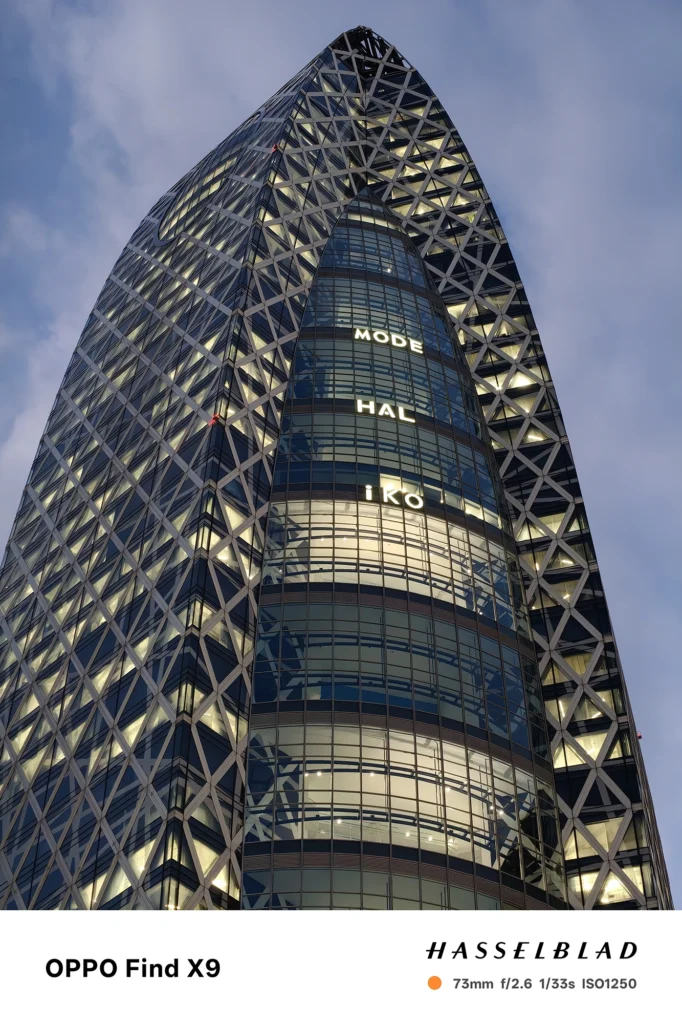 Find X9／カメラ：望遠／焦点距離：73mm相当／内容：格子状外装の高層ビルを望遠で引き寄せ、薄暮の空と窓明かりを分離して圧縮効果の立体感を強調した夜景