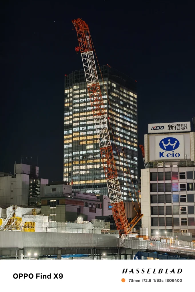 端末名：Find X9／カメラ：望遠／焦点距離：73mm相当／内容：夜空に伸びるクレーンと高層ビル群、駅の看板を望遠で引き寄せ、点光源の反復を圧縮効果で整理した夜景