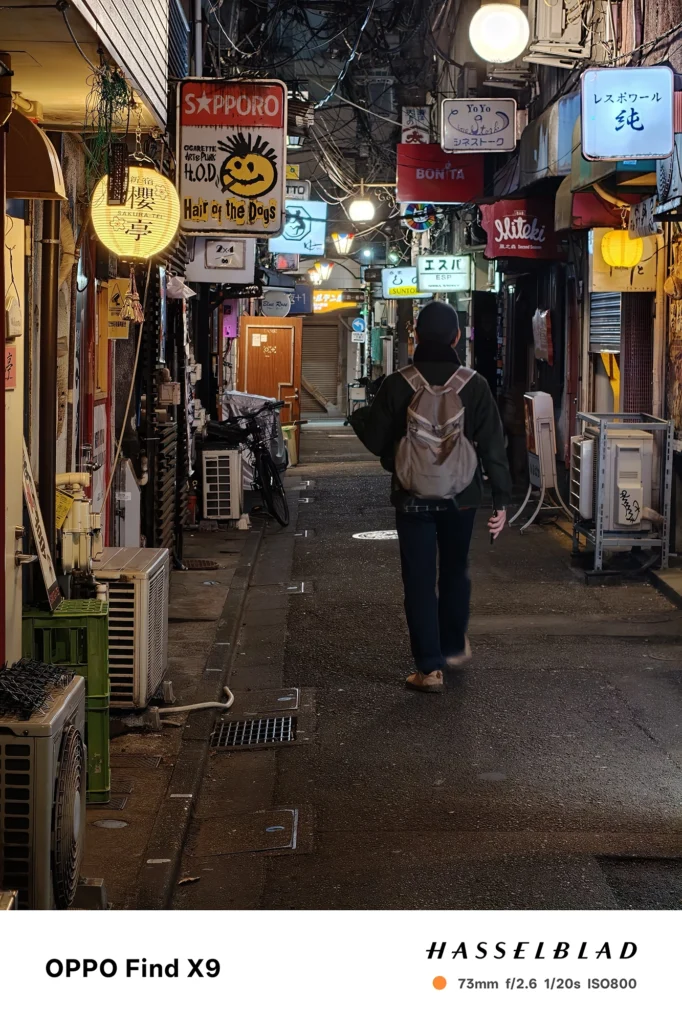 端末名：Find X9／カメラ：望遠／焦点距離：73mm相当／内容：看板と街灯が続く路地を望遠で引き寄せ、歩行者と光源の距離感を圧縮効果でまとめた望遠夜景
