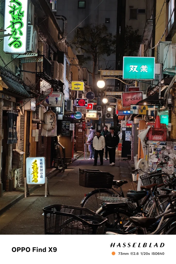 端末名：Find X9／カメラ：望遠／焦点距離：73mm相当／内容：ネオン看板と街灯が密集する路地を望遠で圧縮し、人物の往来と光源の滲みを含めて繁華街の夜景感を強調