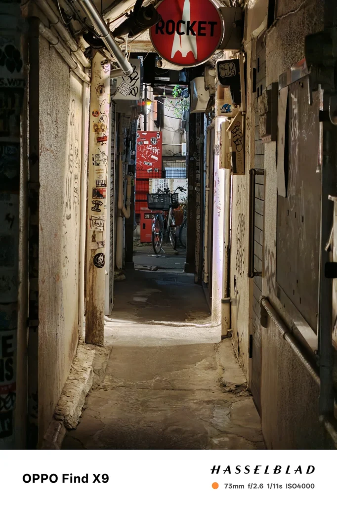 端末名：Find X9／カメラ：望遠／焦点距離：73mm相当／内容：狭い通路の奥行きを望遠で引き寄せ、壁面の落書きや自転車周りの細部を圧縮効果で密度高く見せた夜景