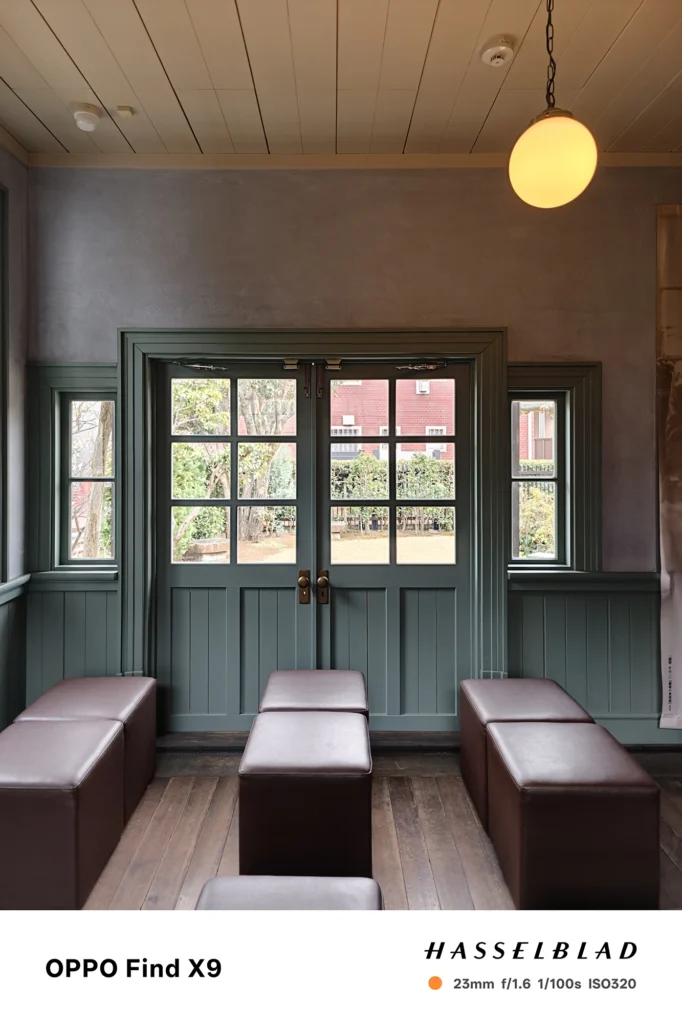 端末名：OPPO Find X9／カメラ：メイン（広角）／焦点距離：23mm相当／内容：木床とベンチのある室内で、格子窓付きの両開き扉から庭の緑が見える逆光気味の内装