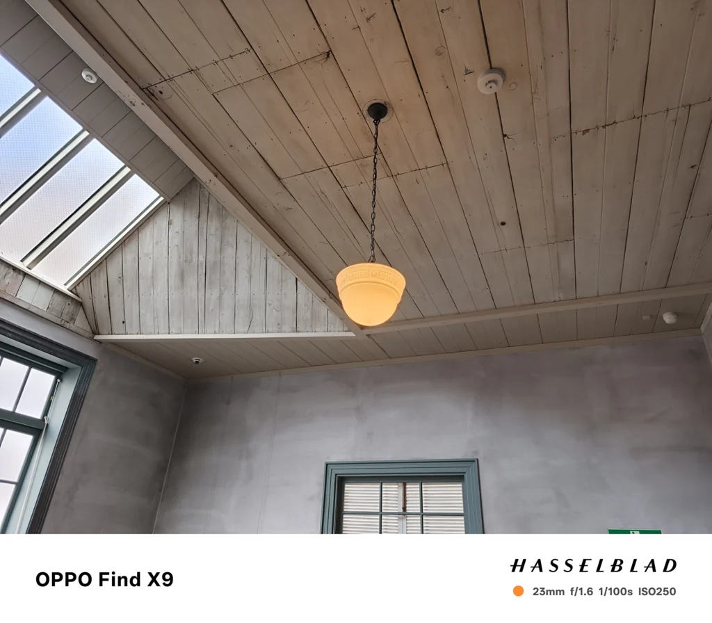 端末名：OPPO Find X9／カメラ：メイン（広角）／焦点距離：23mm相当／内容：木板天井と吊り下げ照明を中心に、左上の天窓から外光が入る室内上向き構図で壁面が広く写る