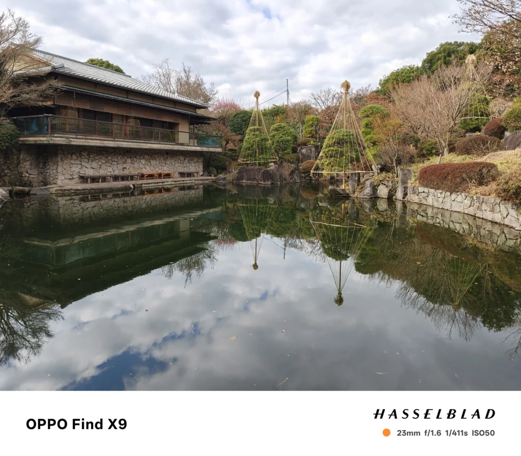 端末名：OPPO Find X9／カメラ：メイン（広角）／焦点距離：23mm相当／内容：池に映る雲と庭木、石垣沿いの日本家屋や雪吊りを広く収めた昼間の日本庭園と水面反射