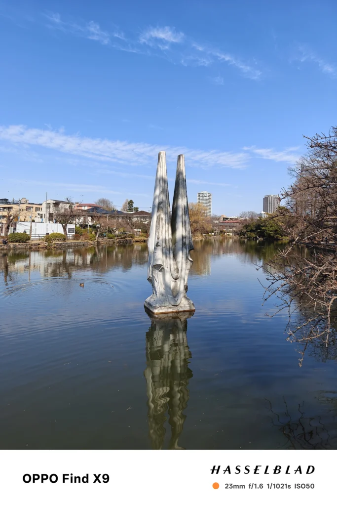 OPPO Find X9の23mm広角で撮影した池の彫刻。水面の反射と背景の住宅街を自然な遠近で描写