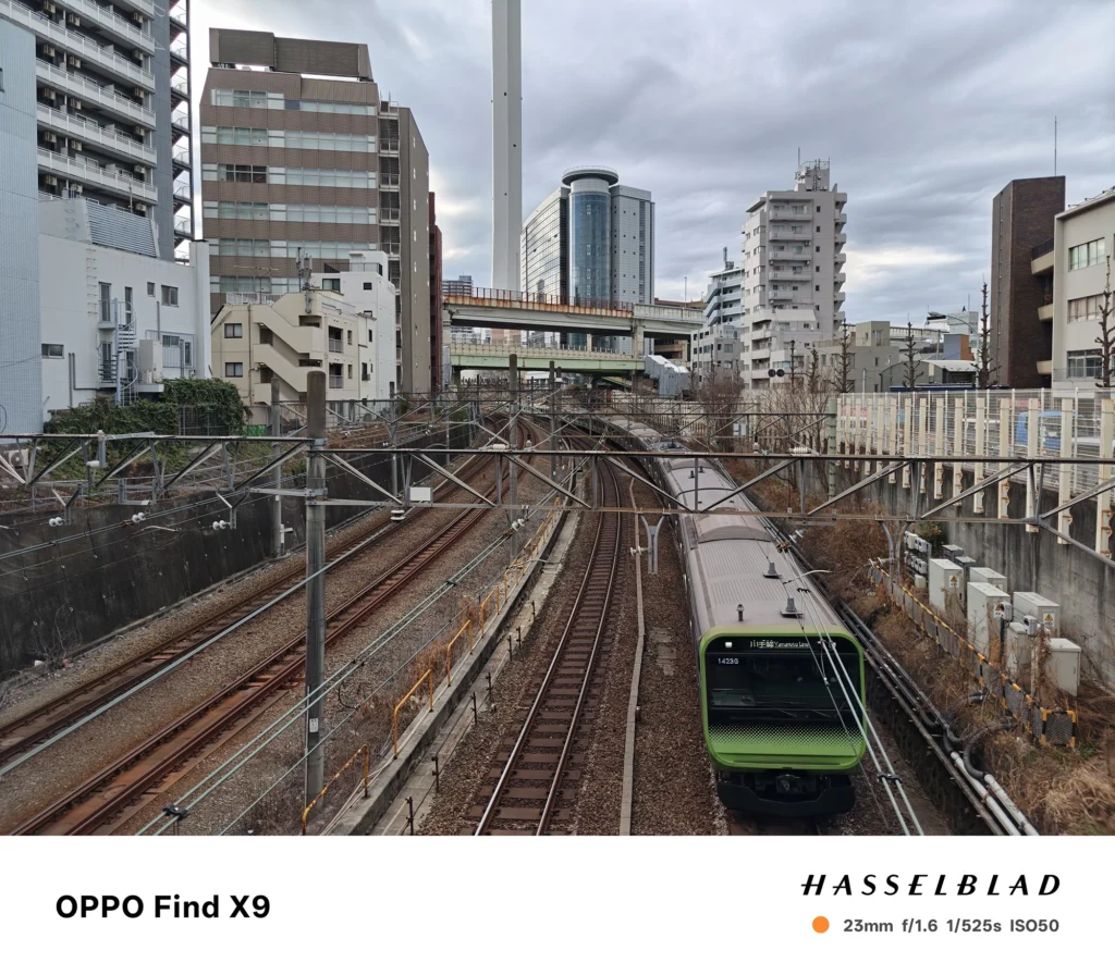 OPPO Find X9／カメラ：メイン（広角）／焦点距離：23mm相当／内容：高架上から見下ろした複数の線路と架線、右手に走る緑の電車、周囲のビル群と曇り空の街並み