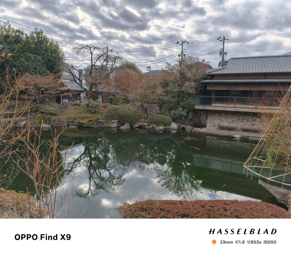 端末名：OPPO Find X9／カメラ：メイン（広角）／焦点距離：23mm相当／内容：池の水面に空と建物が映り、左右に日本家屋と庭木が並ぶ昼間の庭園風景と静かな水面